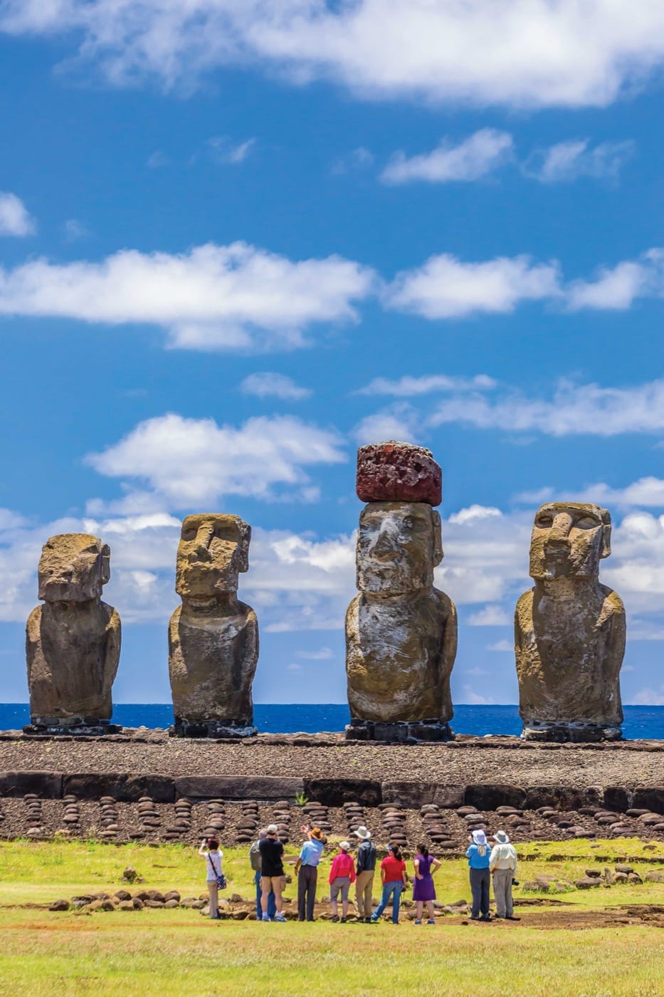 A crowd of people studies a row of great big humanoid stone statues. One of the statues has a large reddish rock balanced on its head